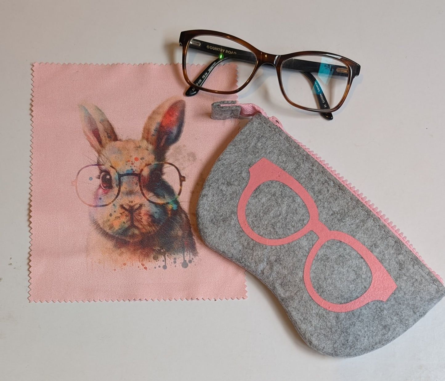 A felt glasses case with a design resembling a cartoon rabbit wearing glasses, displayed next to a pair of glasses and a pink wiping cloth.