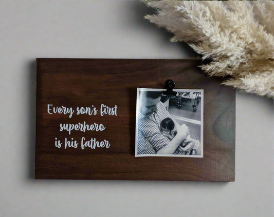 A wooden photo plaque with a black bulldog clip, featuring the text 'Every son's first superhero is his father' in black. A photo of a father and son is attached to the plaque.