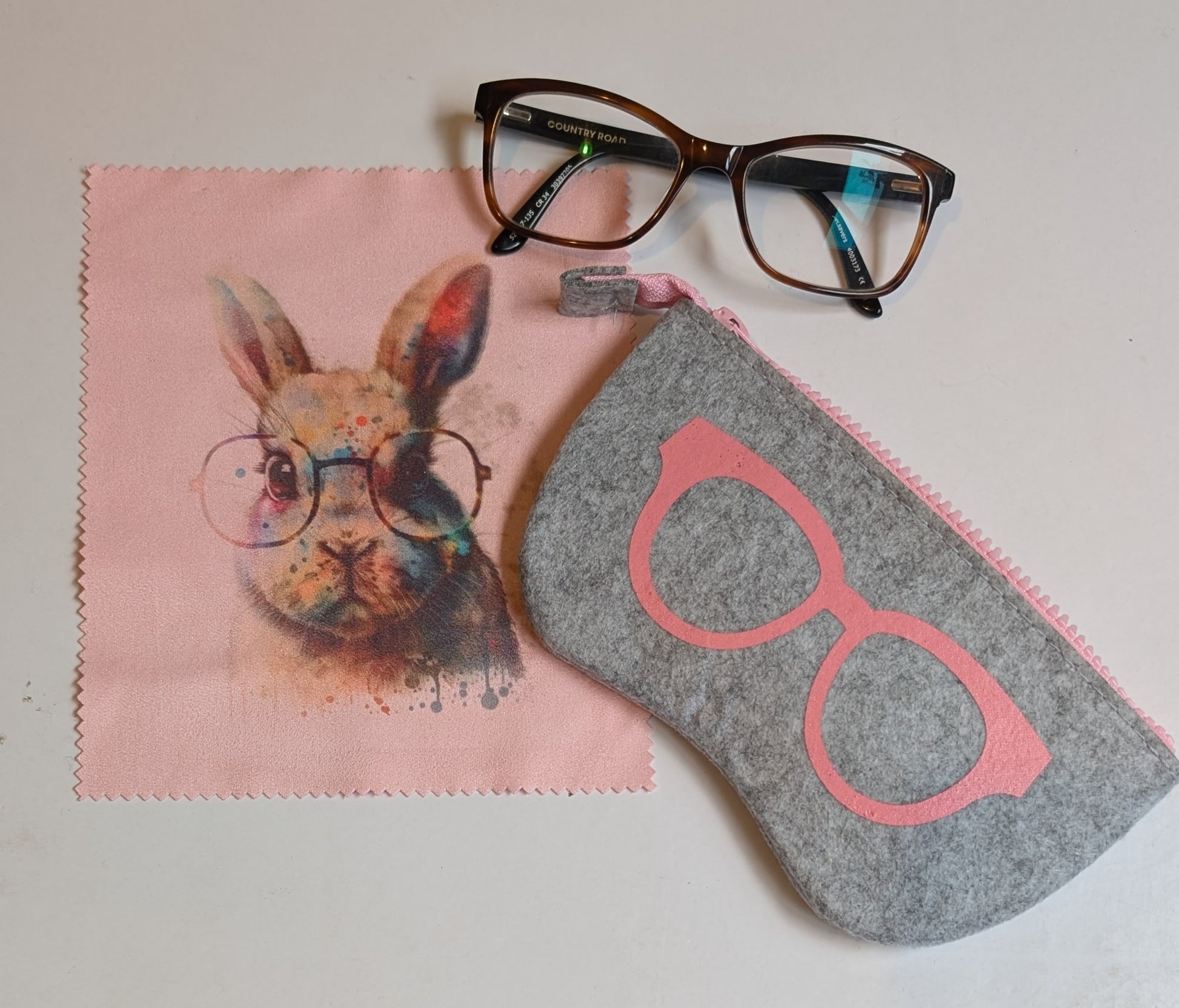 A felt glasses case with a design resembling a cartoon rabbit wearing glasses, displayed next to a pair of glasses and a pink wiping cloth.